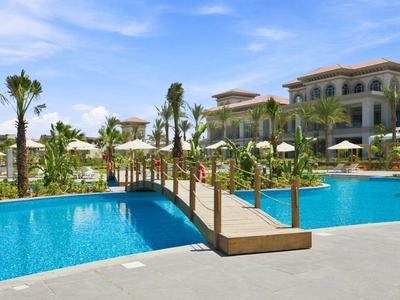 Ein Hotel mit großem Pool, Holzbogenbrücke und Palmen unter blauem Himmel.
