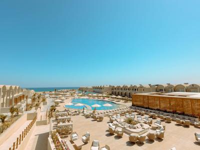 Großer Hotelpool mit runden Liegen, Sonnenschirmen und klar blauem Himmel am Meer im Hintergrund.