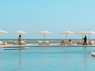 Großer Pool mit blauem Wasser, Liegestühlen und weißen Sonnenschirmen am Meer unter klarem Himmel.