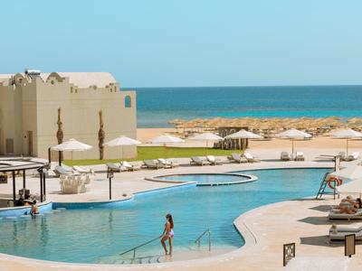 Großer Pool mit Sonnenschirmen, Liegen, paar Personen und Blick aufs Meer bei klarem Himmel.