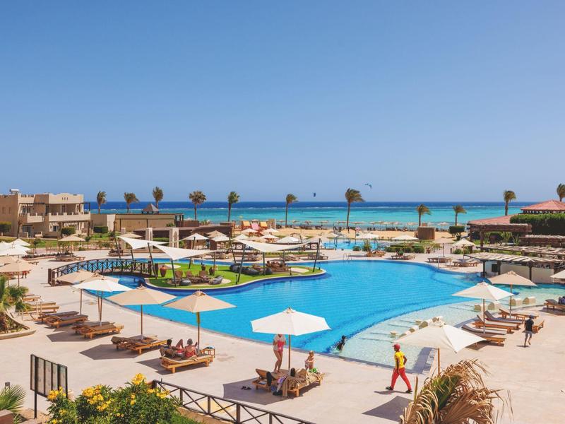 Großer blauer Pool mit Liegestühlen, Sonnenschirmen und Meer im Hintergrund bei sonnigem Himmel.