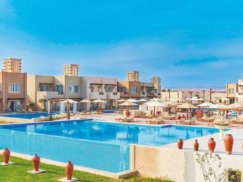 Großer rechteckiger Pool mit Liegestühlen und Sonnenschirmen bei sonnigem Wetter im Hotelkomplex.