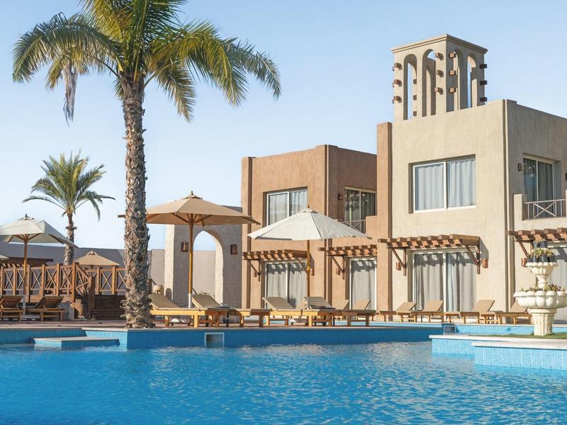 Moderner Gebäudekomplex mit Pool, Palmen und Holzsonnenschirmen unter klarem Himmel.