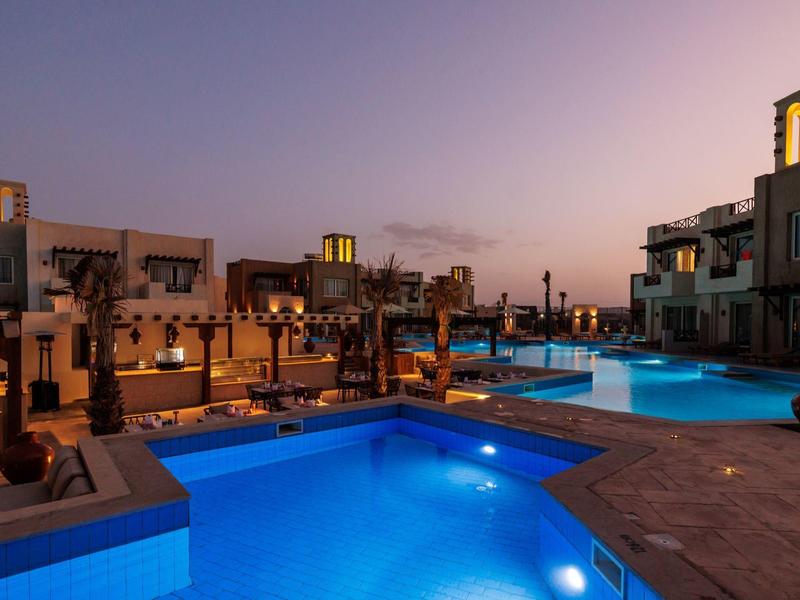 Ein beleuchteter Poolbereich mit mehreren Becken bei Sonnenuntergang, umgeben von modernen Gebäuden.