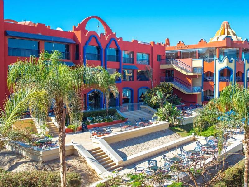 Czerwony hotel z palmami i odkrytym basenem pod błękitnym niebem.