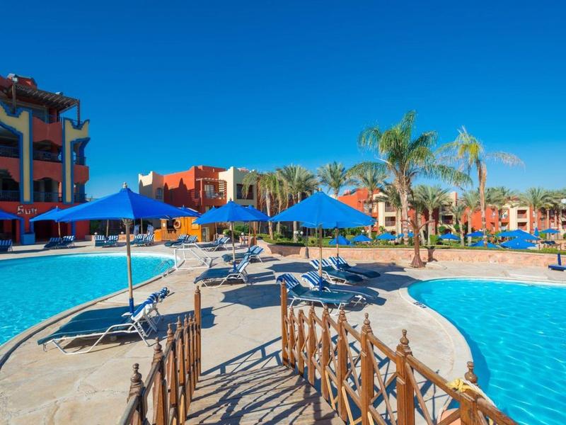 Basen hotelowy z niebieskimi parasolami, leżakami i palmami pod czystym niebem.