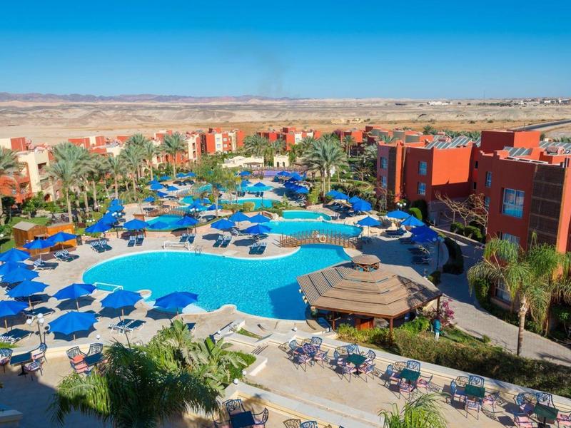 Widok z lotu ptaka na duży ośrodek z basenem i niebieskimi parasolami pod czystym niebem.