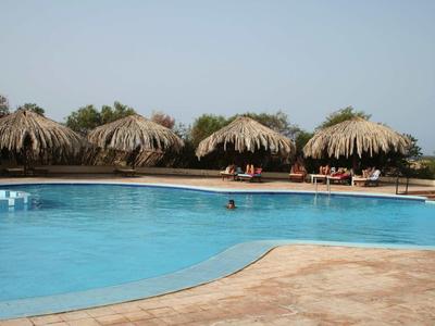 Grande piscina all'aperto con ombrelloni di paglia e aree salotto ai bordi sotto un cielo limpido.