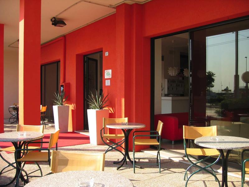 Area terrazza con tavoli, sedie e pareti rosse, vista su giardino e acqua.