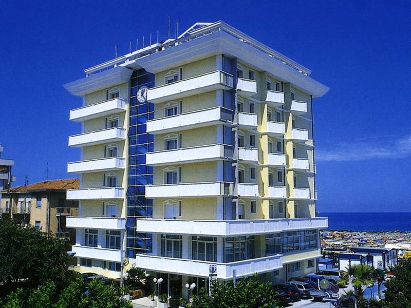 Mehrstöckiges, modernes Gebäude mit gelb-weißer Fassade vor blauem Himmel und Meer.