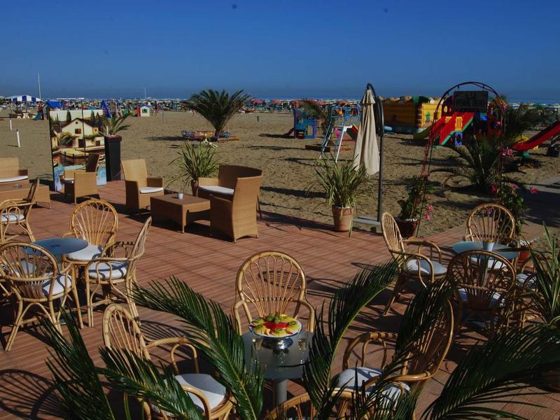 Strand mit Sand, Holzterrasse, Korbstühlen, Tischen, Sonnenschirmen und buntem Spielplatz im Hintergrund.