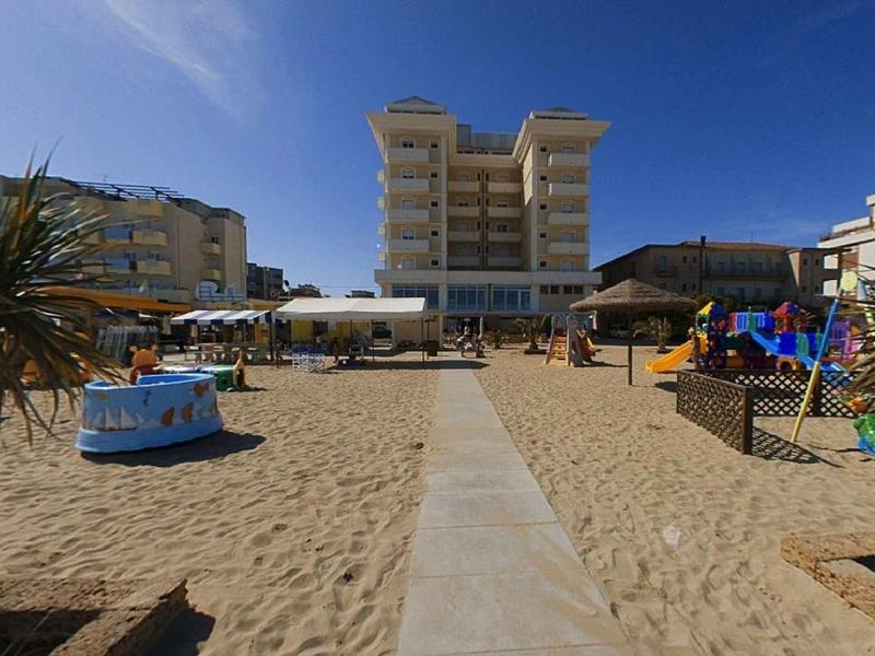 Sandstrand mit Spielplatz, Palmen und mehrstöckigen Gebäuden unter blauem Himmel.