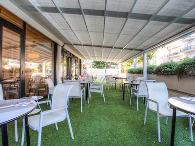 Terrasse mit weißen Stühlen und Tischen auf grünem Kunstrasen unter weißem Sonnendach.