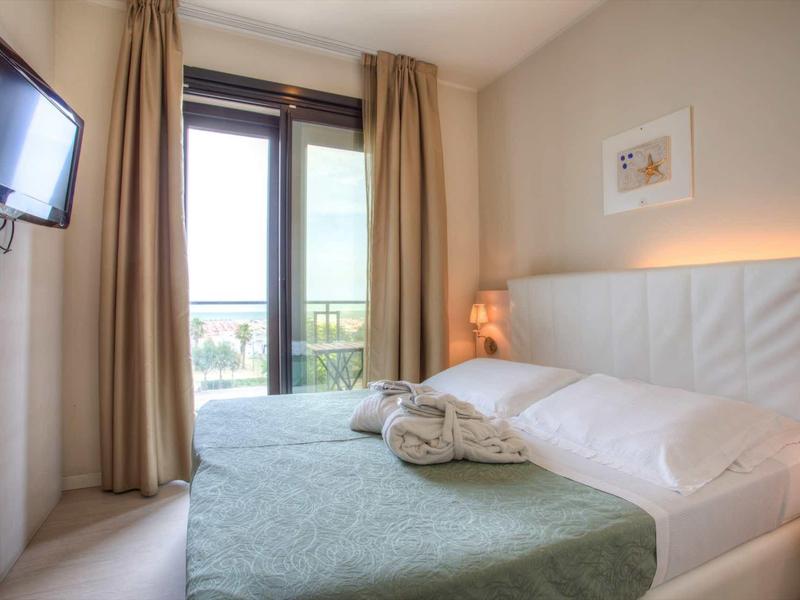 Helles Schlafzimmer mit großem Fenster, weißem Bett, grünem Bettüberwurf und beige Vorhängen.