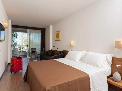 Hell eingerichtetes Hotelzimmer mit Doppelbett, Sofa, kleinem Balkon und rotem Koffer.