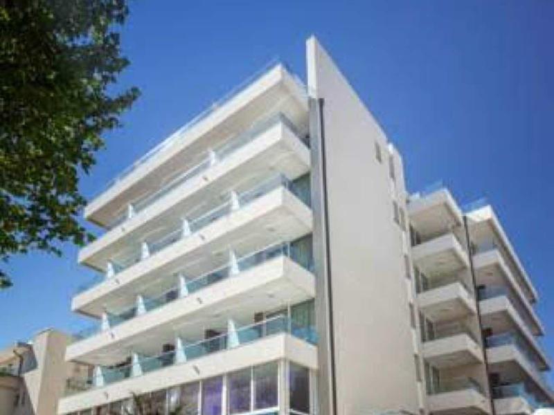 Moderní vícepodlažní hotelová budova s balkony, modrá obloha a stromy v popředí.