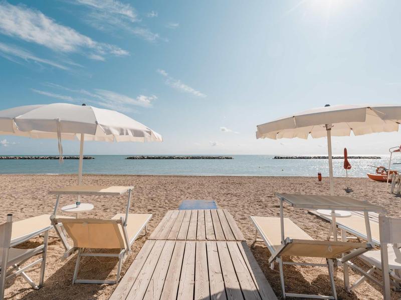 Sandy beach with sun umbrellas, lounge chairs, and wooden walkway leading to the sea under clear sky.