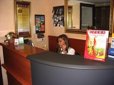 Rezeption mit dunkelgrauem Tresen, Frau am Telefon und Plakaten an der Wand in warmem Raum.