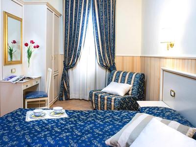 Kleines, helles Zimmer mit blau gemusterter Bettdecke, Sessel, Vorhängen und Wandschrank.