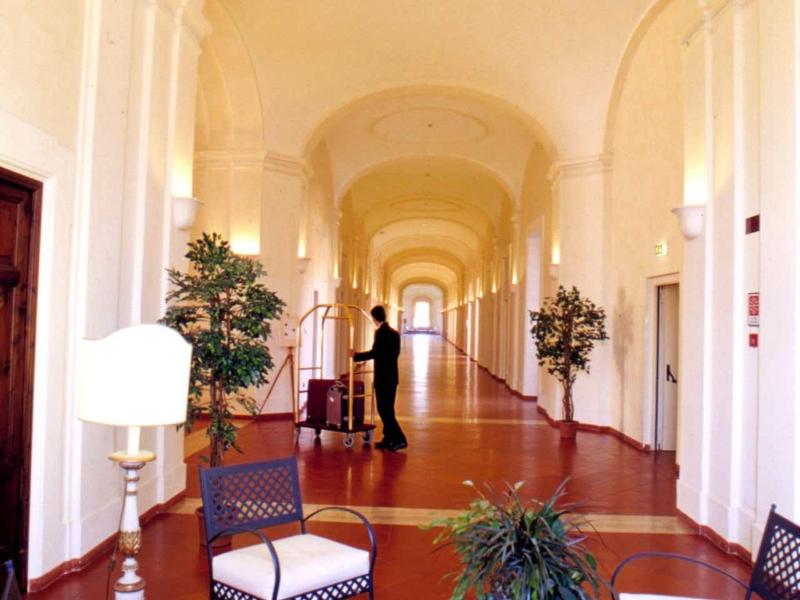 Langer Gang mit gewölbter Decke, weißen Wänden, Holzboden, Sitzgruppen und Pflanzen in Töpfen.