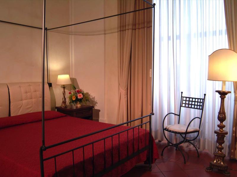 Schlafzimmer mit roten Bettdecke, schwarzem Metallbett und Stehlampe neben einem Fenster mit grauen Gardinen.