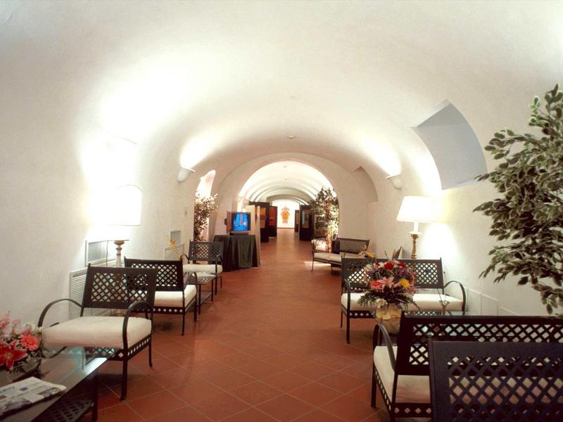 Langer, heller Gewölbegang mit weißen Wänden, Stühlen, Tischen und Pflanzen an den Seiten.