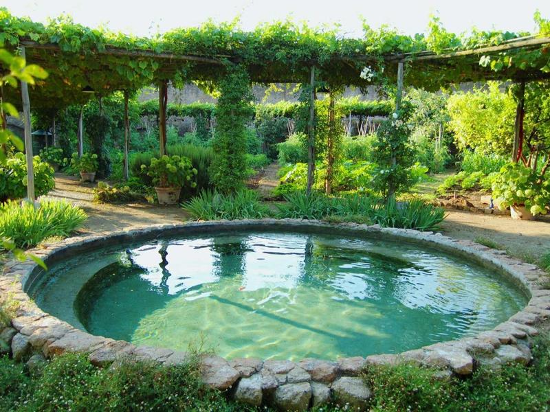 Rundes Wasserbecken mit Steinumrandung, umgeben von grünem Garten und bepflanztem Pergola-Bogen.