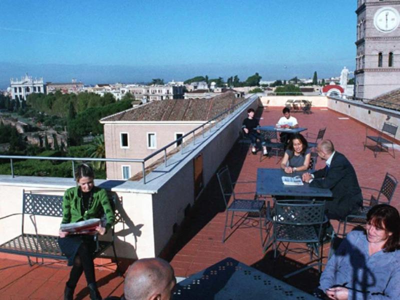 Menschen sitzen an Tischen auf einer sonnigen Dachterrasse mit Blick auf Gebäude und blauen Himmel.