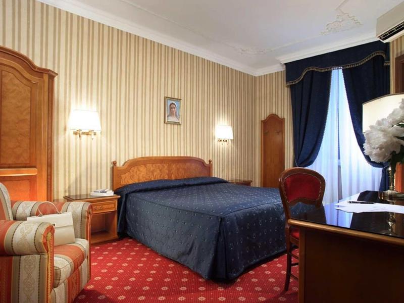 Classic hotel room with double bed, two nightstands, armchair, and desk under blue curtain.