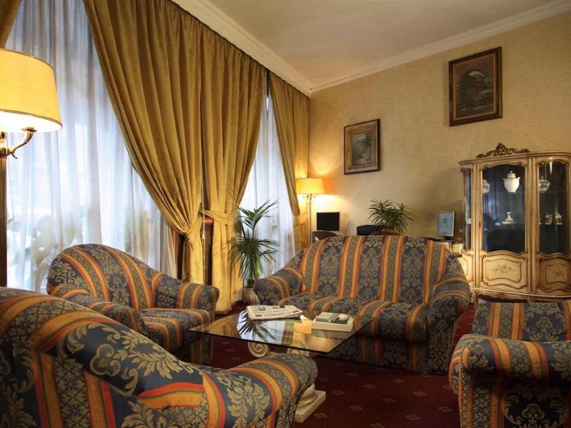 Cozy hotel lounge with upholstered armchairs, sofa, lamp, and curtains in warm tones.