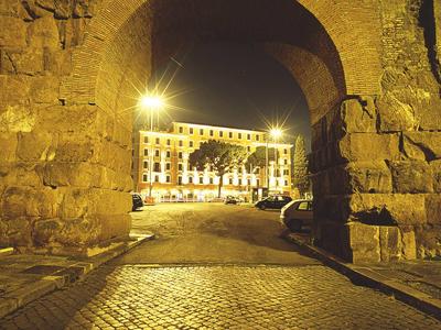 Großer Steinbogen mit Kopfsteinpflaster beleuchtet, dahinter Gebäude und Laternen bei Nacht.