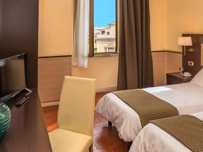 Hotel room with two single beds, TV, chair, and window overlooking buildings.