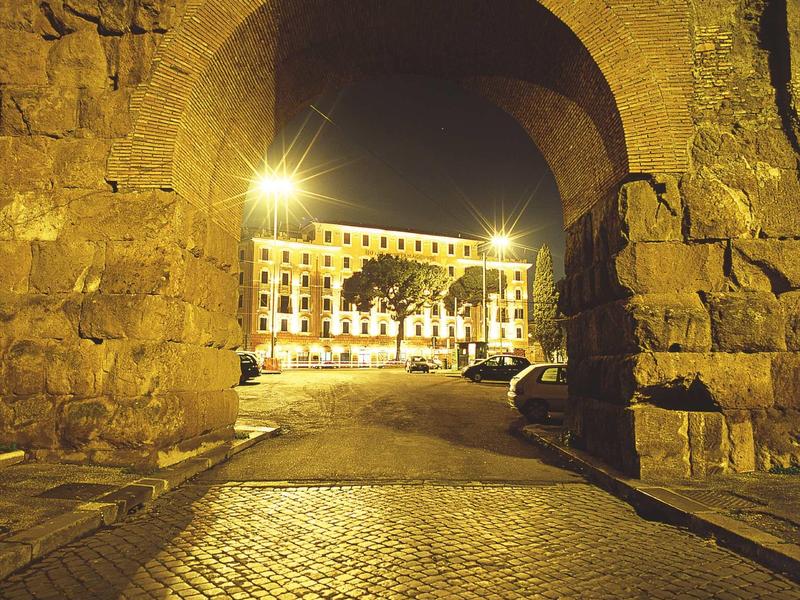 Illuminated cobblestone path leads through stone archway to historic building at night.
