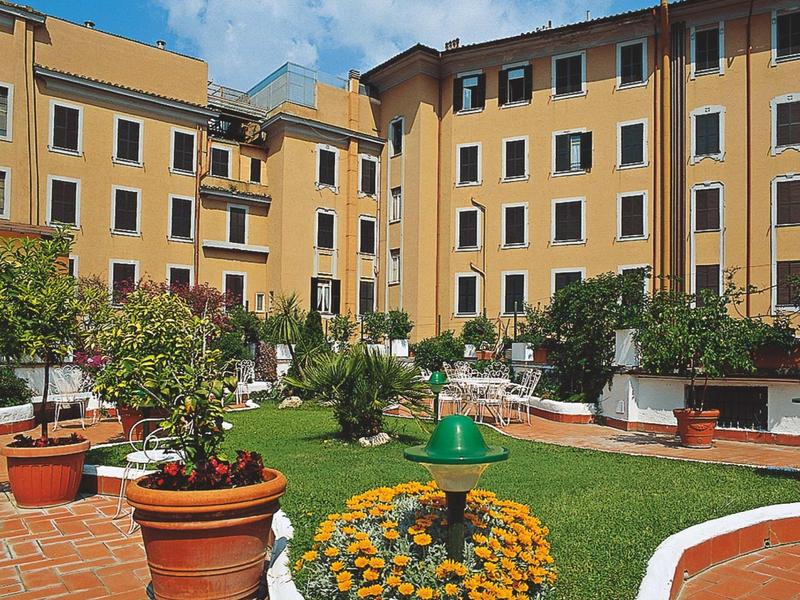 Sunny garden area with flowers, pots, and seating in front of a yellow apartment building.