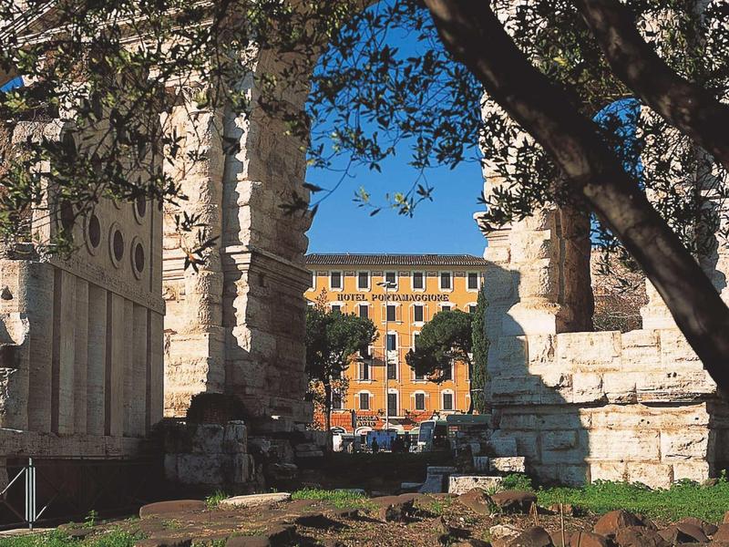 Ancient ruins with a large stone arch and trees in the foreground, city buildings in the background.