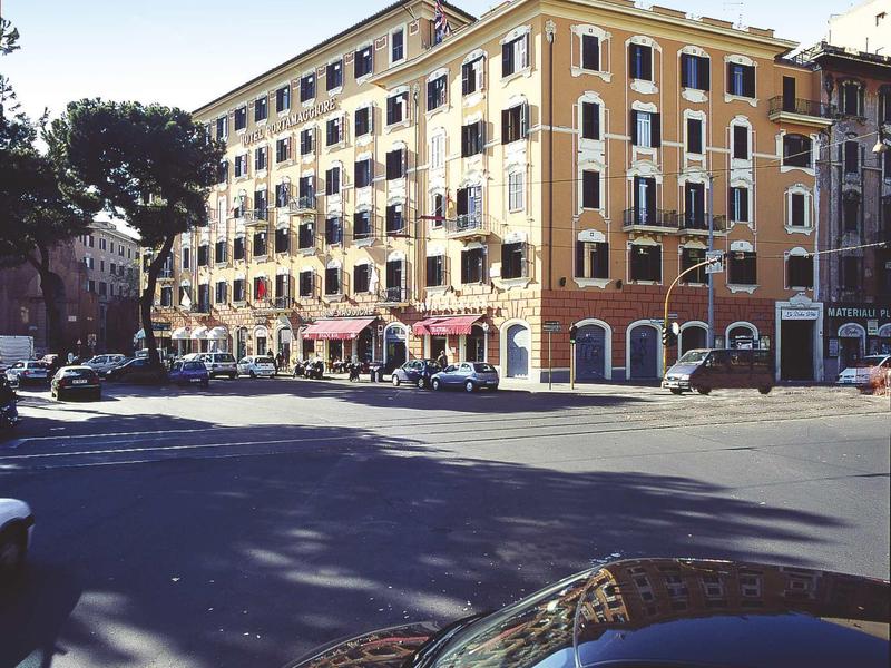 Large hotel building on a sunlit street corner with parked cars.