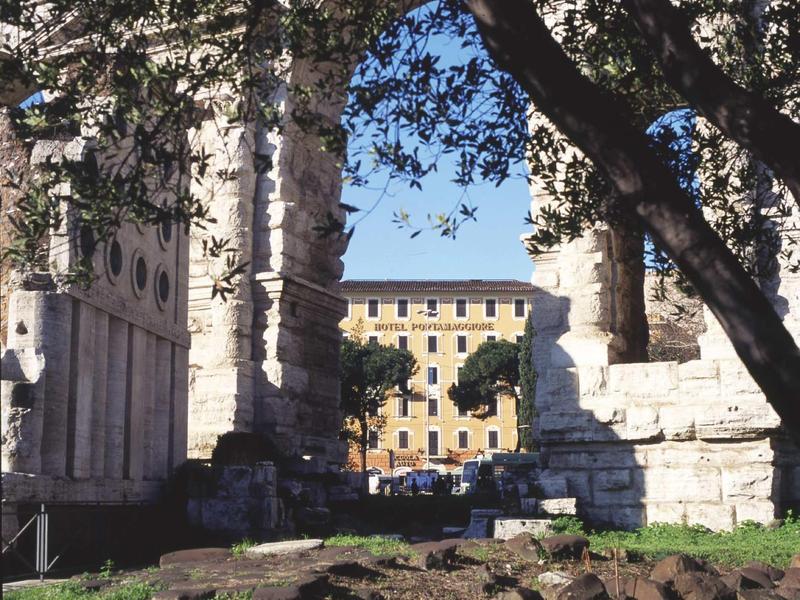 Historic stone ruins with trees and a yellow building in the background under clear sky.