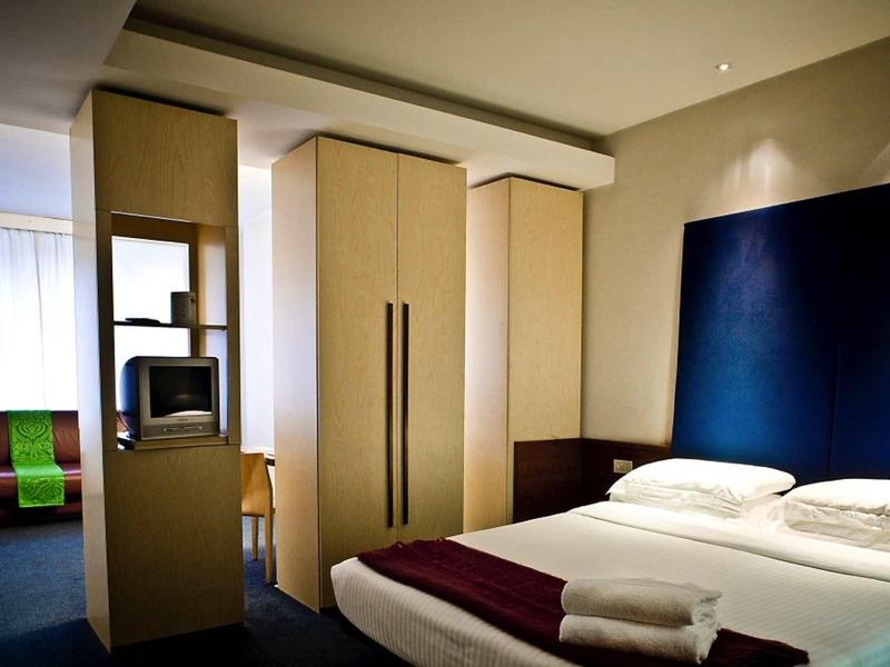 Modern eingerichtetes Hotelzimmer mit großem Bett, Fernseher und Kleiderschrank, helle Farben.