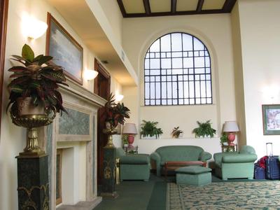 Světlá hotelová hala se zelenými křesly, velkými rostlinami a krbem pod vysokým oknem.