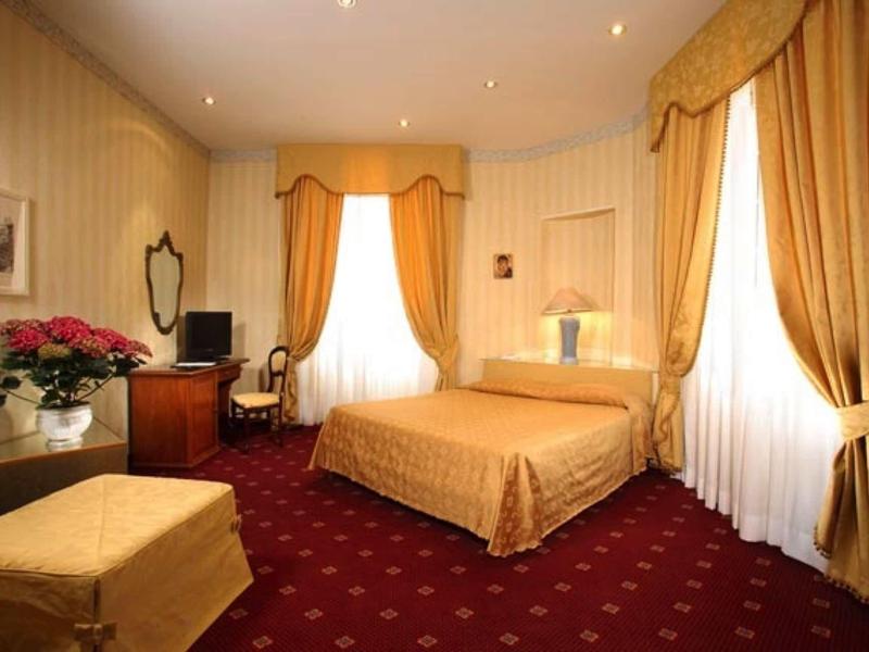 Elegantní hotelový pokoj s manželskou postelí, červeným kobercem a zlatými záclonami.