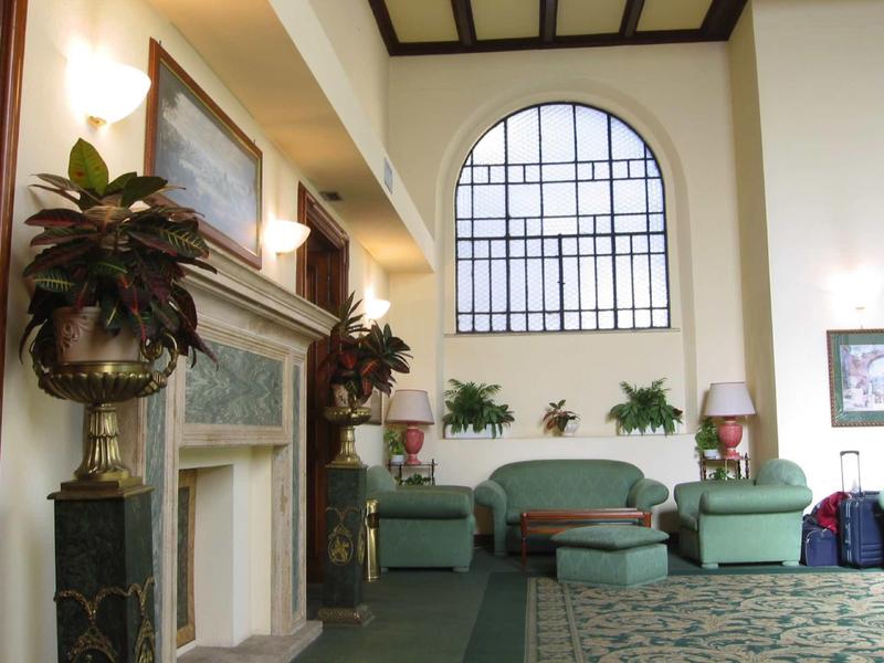 Světlá hotelová hala se zelenými křesly, velkými rostlinami a krbem pod vysokým oknem.