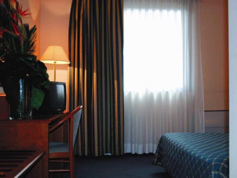 Kleines Hotelzimmer mit Tür, Fenster mit Vorhängen, Fernseher, Schreibtisch und einem Bett.