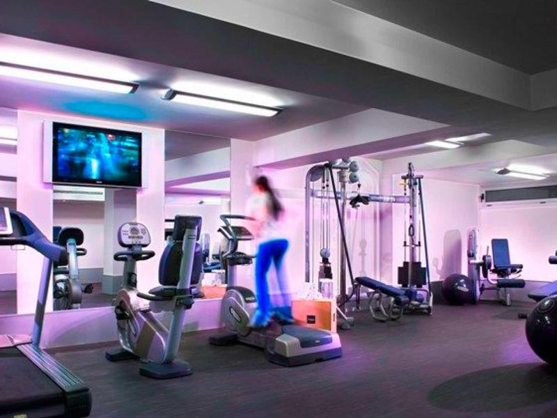 Fitnessraum mit Trainingsgeräten, voller bunter Beleuchtung und einer Person im Sportoutfit.