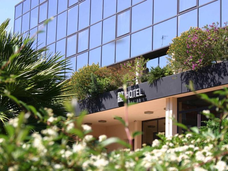 Modernes Hotelgebäude mit Glasfassade, umgeben von grünen Pflanzen und bunten Blumen.