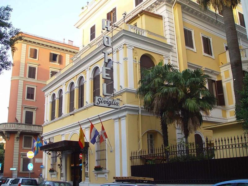 Geel meerlaagse gebouw met vlaggen en palmbomen bij de ingang van een hotel.