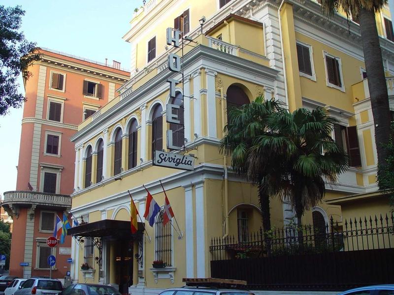 Geel historisch hotelgebouw met vlaggen en palmbomen bij de ingang.