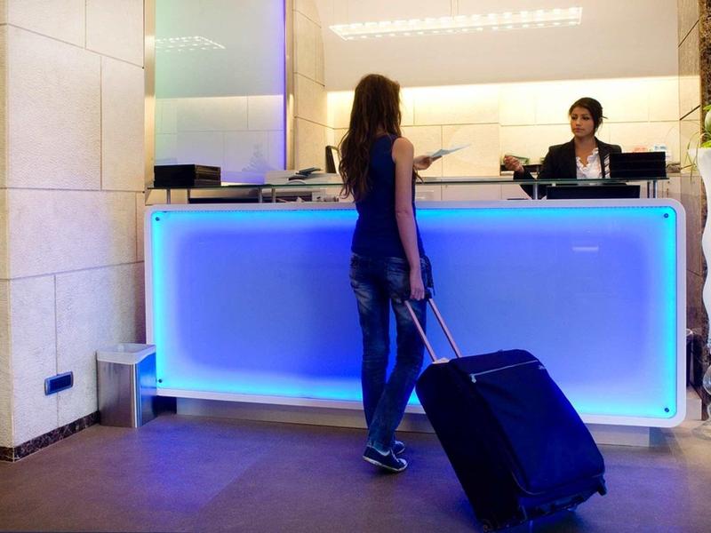 Eine Frau mit Rollkoffer steht am beleuchteten Empfangstresen eines Hotels.