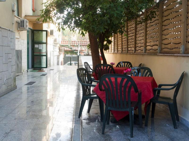 Venkovní prostor s červeně pokrytým stolem a černými židlemi pod stromem vedle zdi