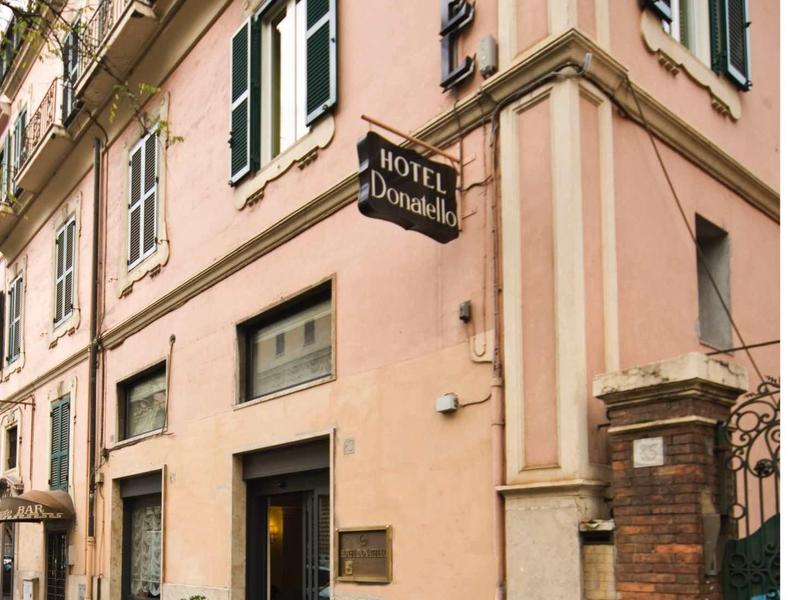 Eckgebäude in Hellrosa mit grün-weißen Fensterläden und Hotelschildern.