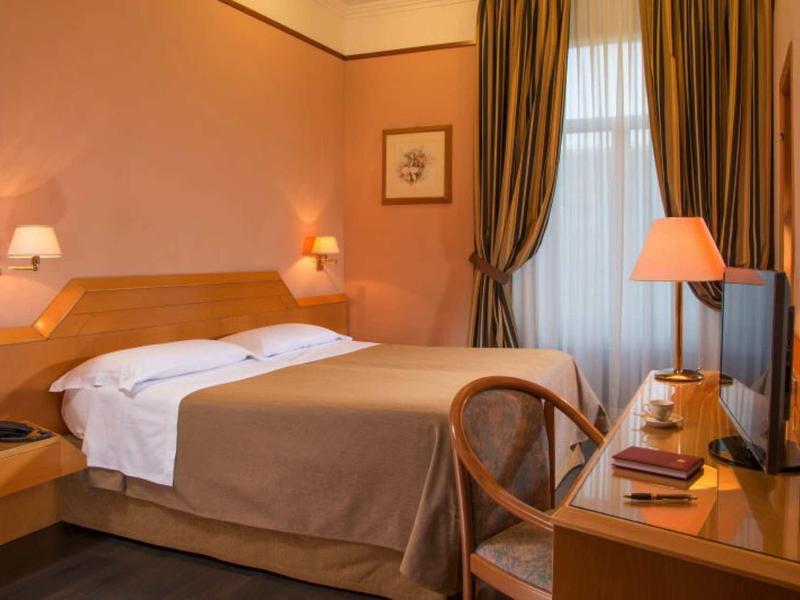 Gemütliches Hotelzimmer mit Doppelbett, Holzmöbeln, Tischlampe und braunen Vorhängen.